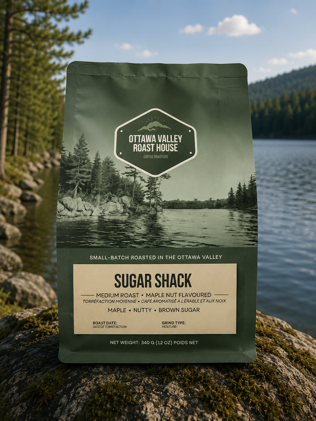 A stand-up pouch of Ottawa Valley Roast House Sugar Shack coffee stands on a mossy rock with a lake and forested mountains in the background. The pouch is dark olive green with a beige label panel and a scenic forest-and-lake photo on the front.