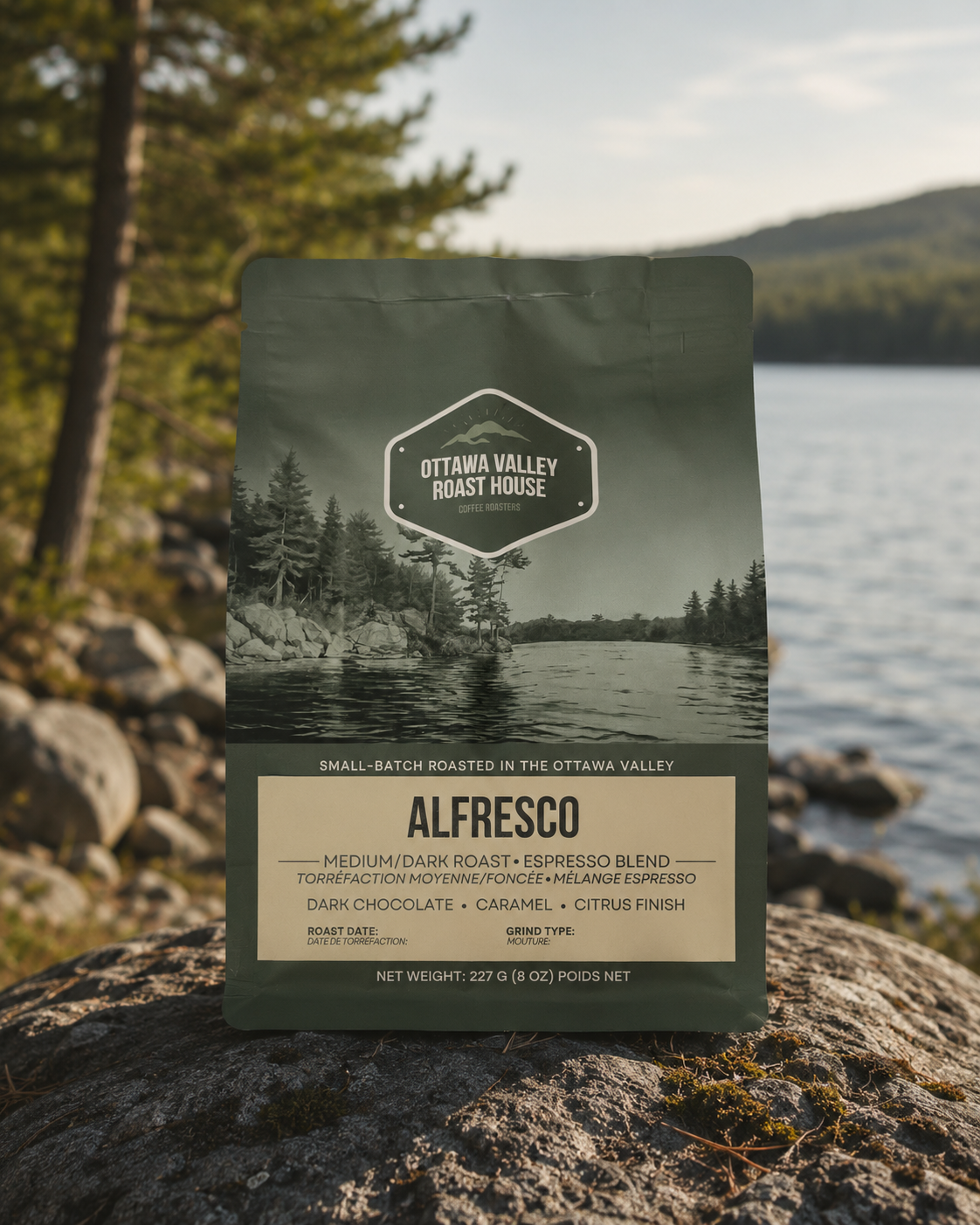 A bag of Ottawa Valley Roast House Alfresco coffee stands upright on a rock with a lake and forest in the background. The package is dark olive green with a scenic lake-and-trees label and a beige lower panel showing the product name.