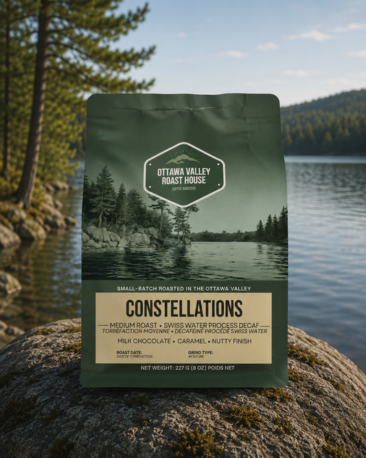 A bag of Ottawa Valley Roast House Constellations coffee stands on a large rock in front of a lake and forest landscape. The package is dark green with a scenic label and a cream-colored bottom panel.