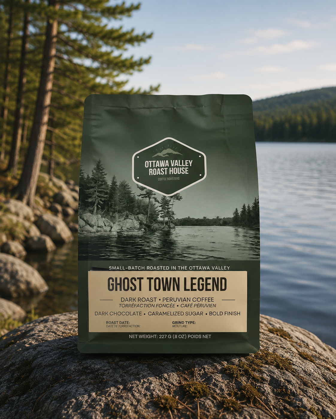 A bag of Ottawa Valley Roast House Ghost Town Legend coffee stands on a large rock in front of a lake and pine forest. The bag is dark green with a scenic lake-and-mountain label and a cream-colored lower label panel.