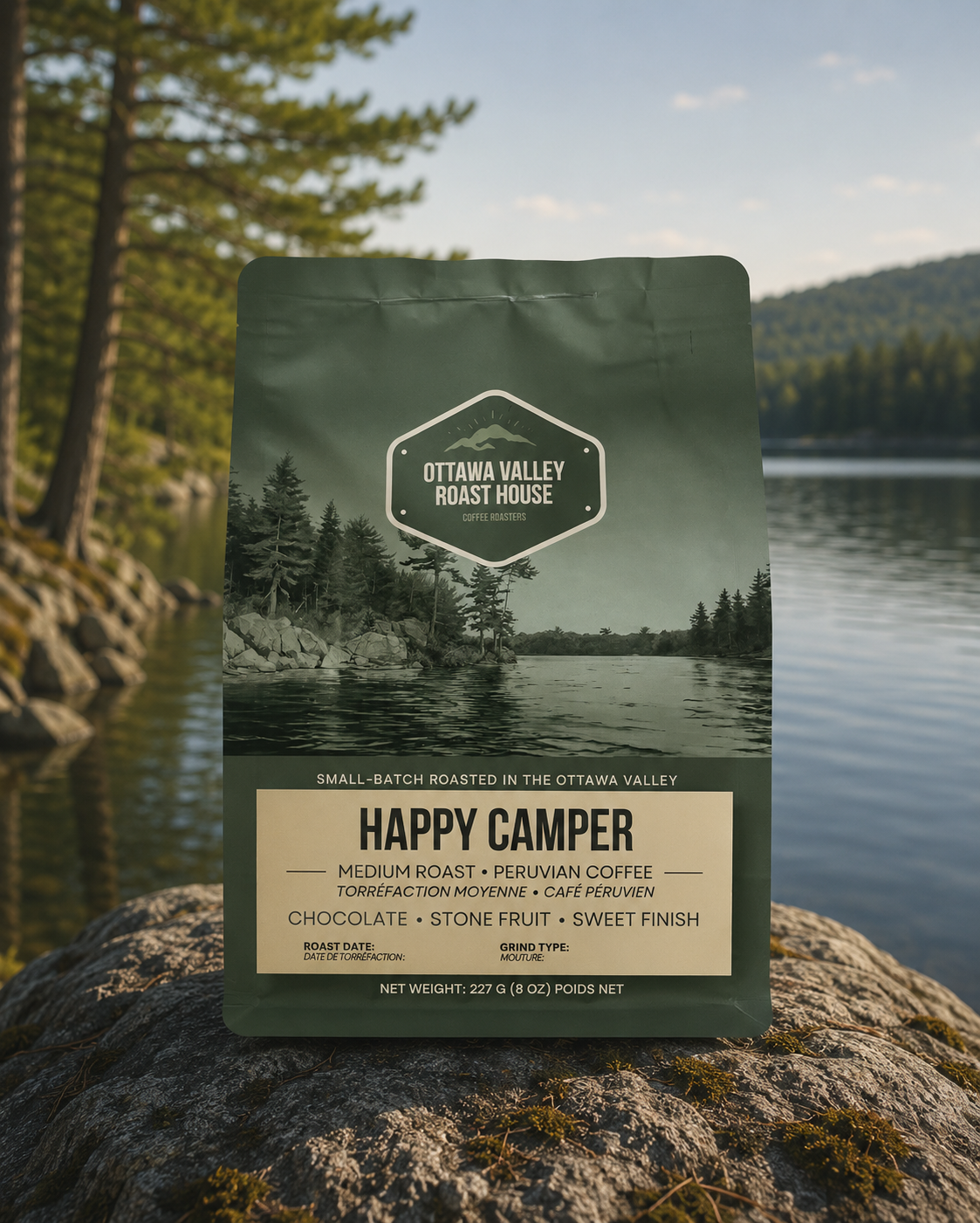 A bag of Ottawa Valley Roast House Happy Camper coffee stands upright on a large rock with a blurred lake and forest landscape in the background.