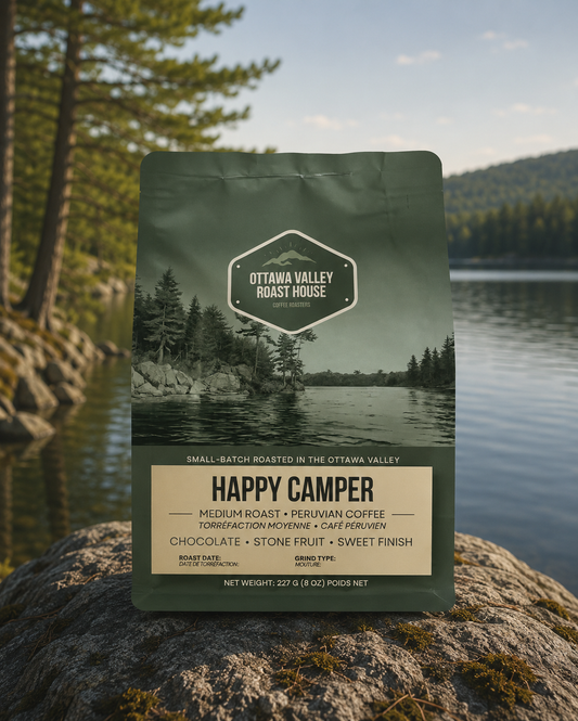 A bag of Ottawa Valley Roast House Happy Camper coffee stands upright on a large rock with a blurred lake and forest landscape in the background.
