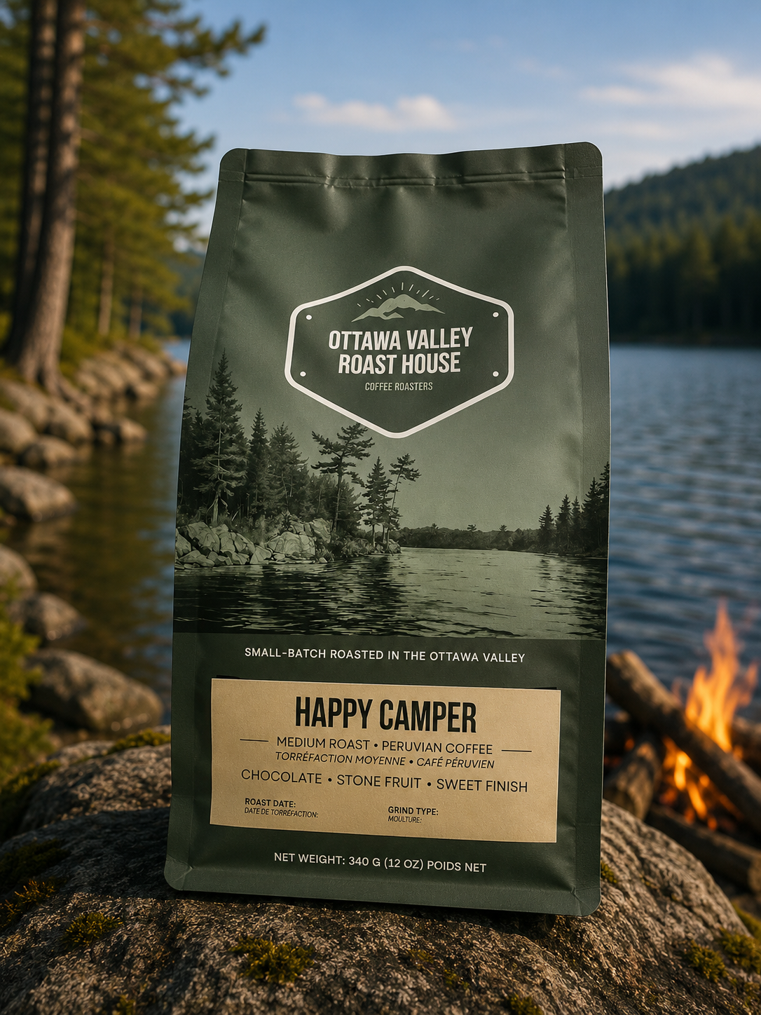 A bag of Ottawa Valley Roast House Happy Camper coffee stands upright on a rock outdoors by a lake, with trees, water, and a small fire visible in the background.