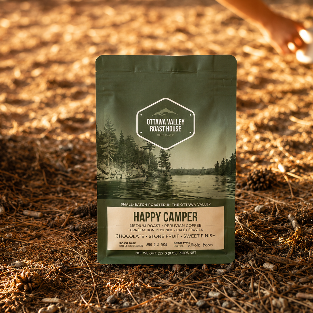 A bag of Ottawa Valley Roast House 'Happy Camper' coffee stands upright on a forest floor covered in pine needles and dry leaves, with a person's arm and leg blurred in the background near a tree trunk.