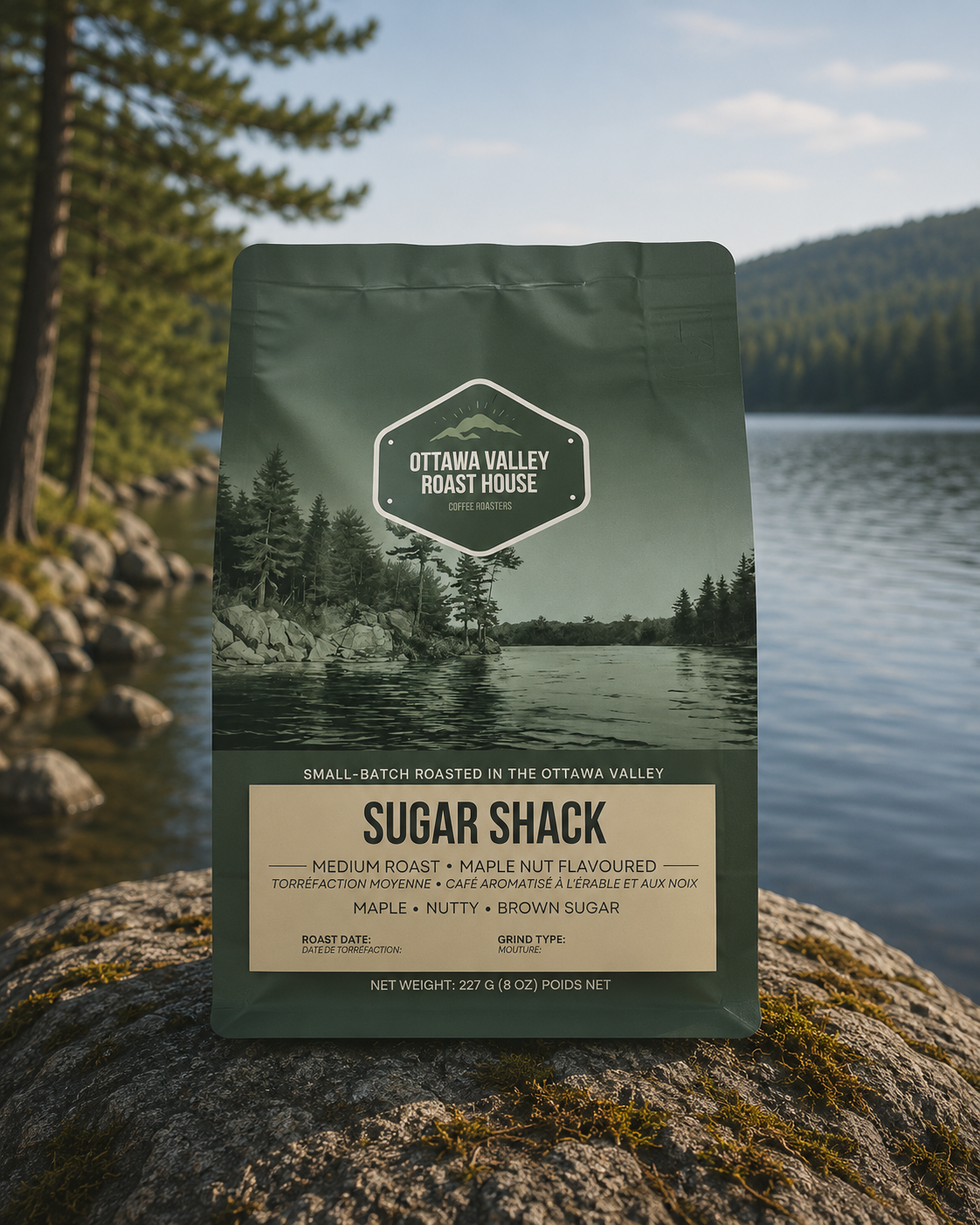 A bag of Ottawa Valley Roast House Sugar Shack coffee stands upright on a mossy rock with a lake and forest landscape in the background.