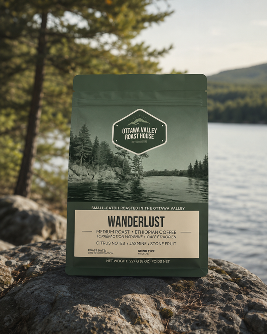 A bag of Ottawa Valley Roast House Wanderlust coffee stands upright on a large rock with a blurred outdoor lakeside and forest background.
