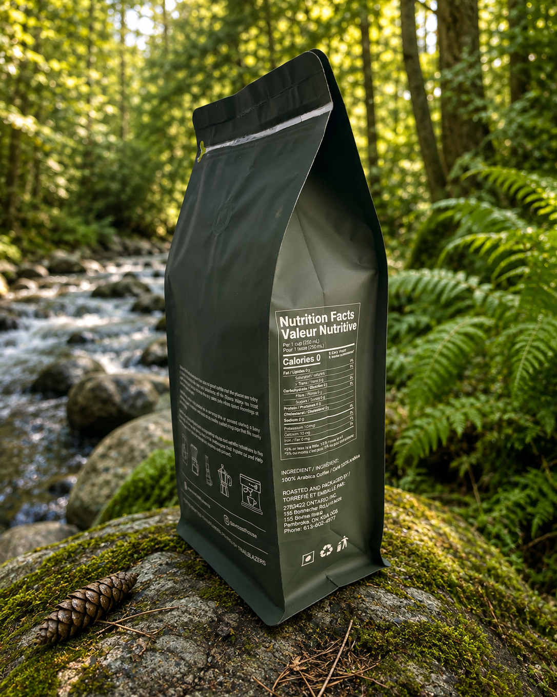 A matte black resealable pouch of nutrition supplement or powder stands on a mossy rock in a forest with a stream and ferns in the background. The front label shows 'Nutrition Facts' and 'Calories 5'.