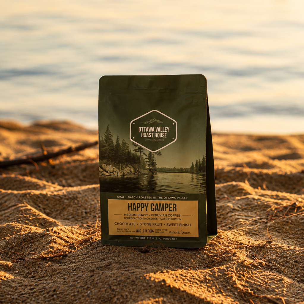 A bag of Ottawa Valley Coffee Roasters Happy Camper coffee stands upright on a sandy shore with a lake and sunset in the background.