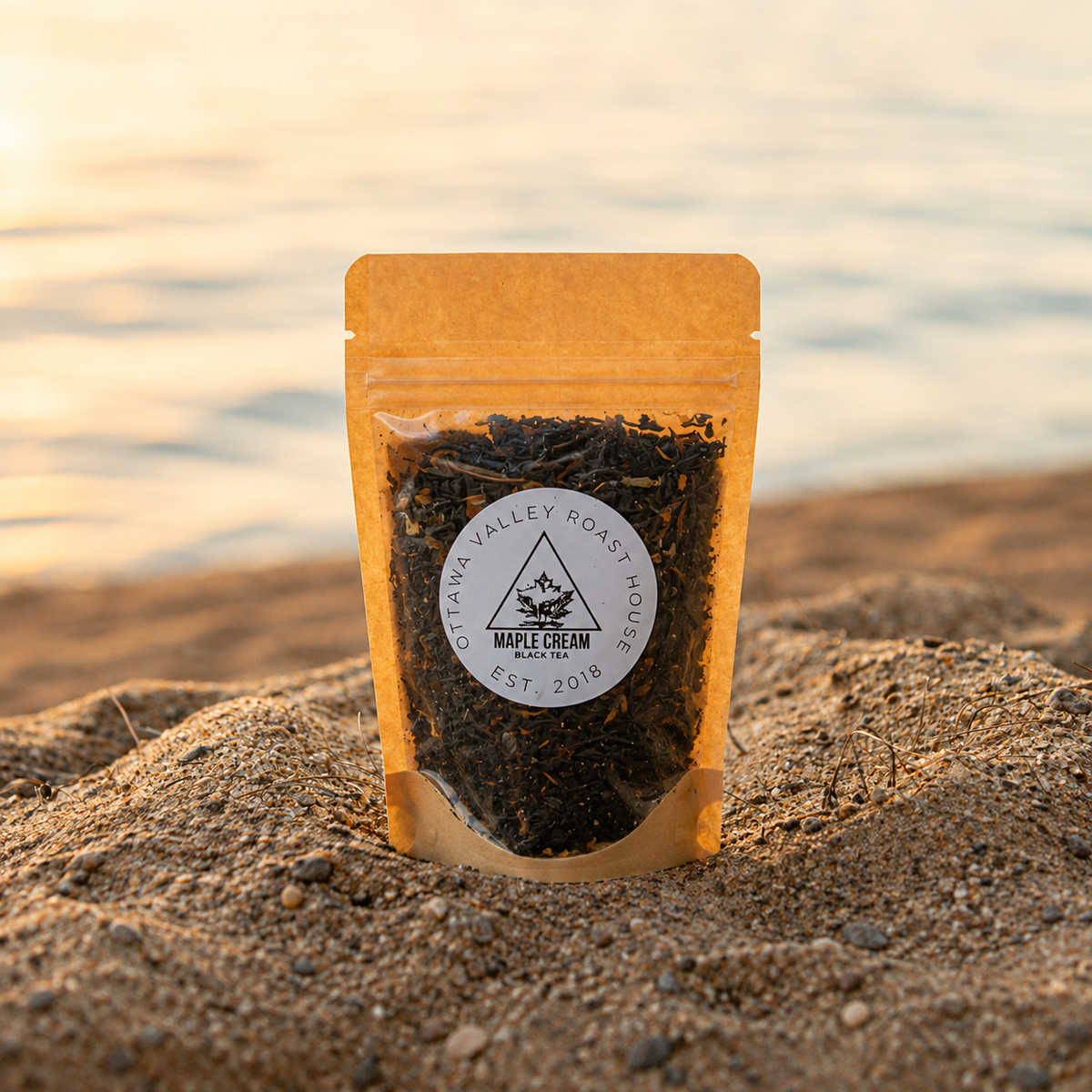 A stand-up kraft paper pouch of Maple Valley Roast Coffee Tea by Star Lake Coffee Roasters, standing upright in sand with a blurred beach and water background at sunset.