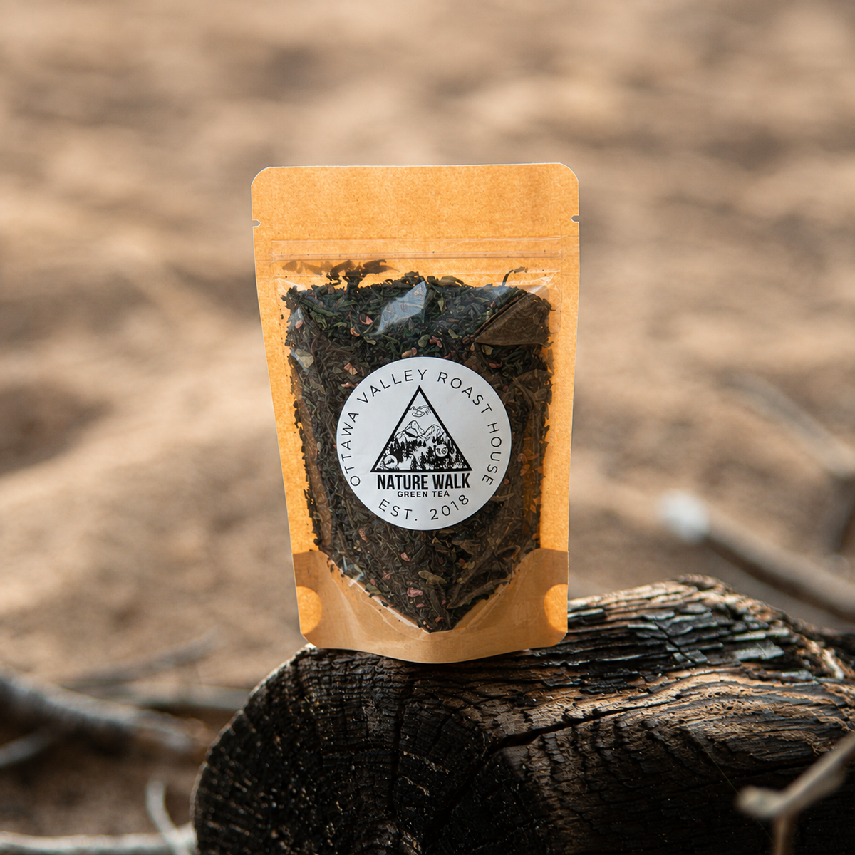 A stand-up pouch of dark roasted coffee leaves standing on a cut tree stump outdoors, with a blurred sandy ground and branches in the background. The pouch is tan with a circular white Nature Walk Coffee Roasters label.