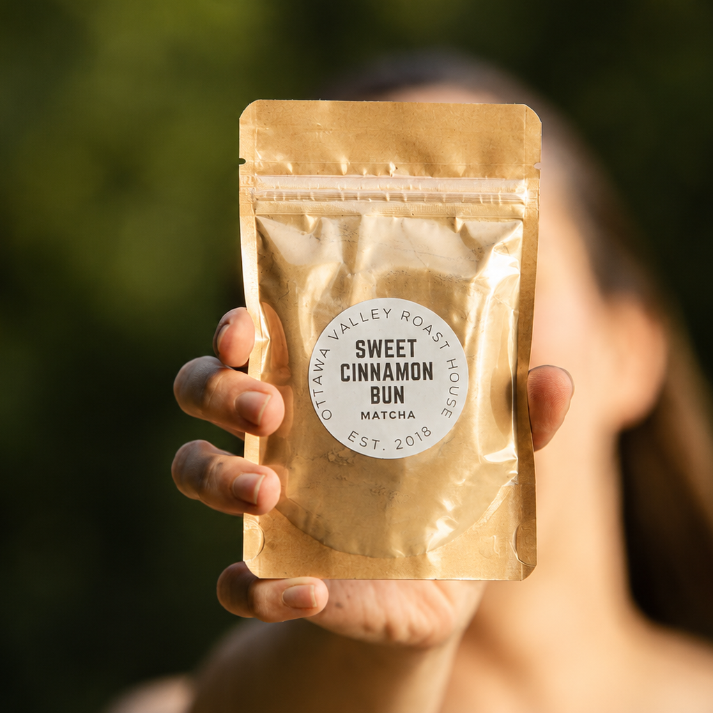 A person holds up a gold-colored stand-up pouch of Sweet Cinnamon Bun Matcha from Alaya Roastery against a blurred green outdoor background.