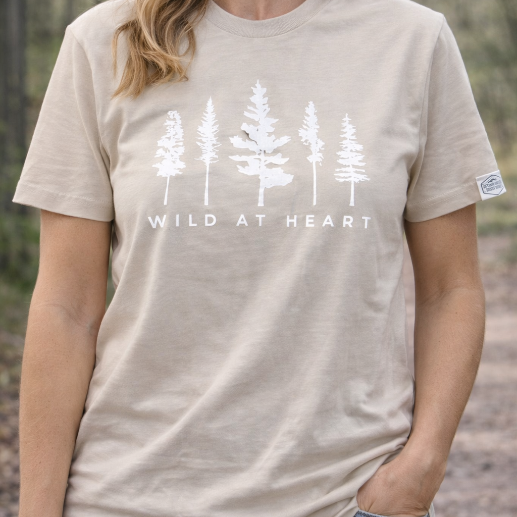 Person wearing a beige t-shirt with white tree design and 'Wild at Heart' text in a forest setting.