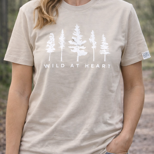 Person wearing a beige t-shirt with white tree design and 'Wild at Heart' text in a forest setting.
